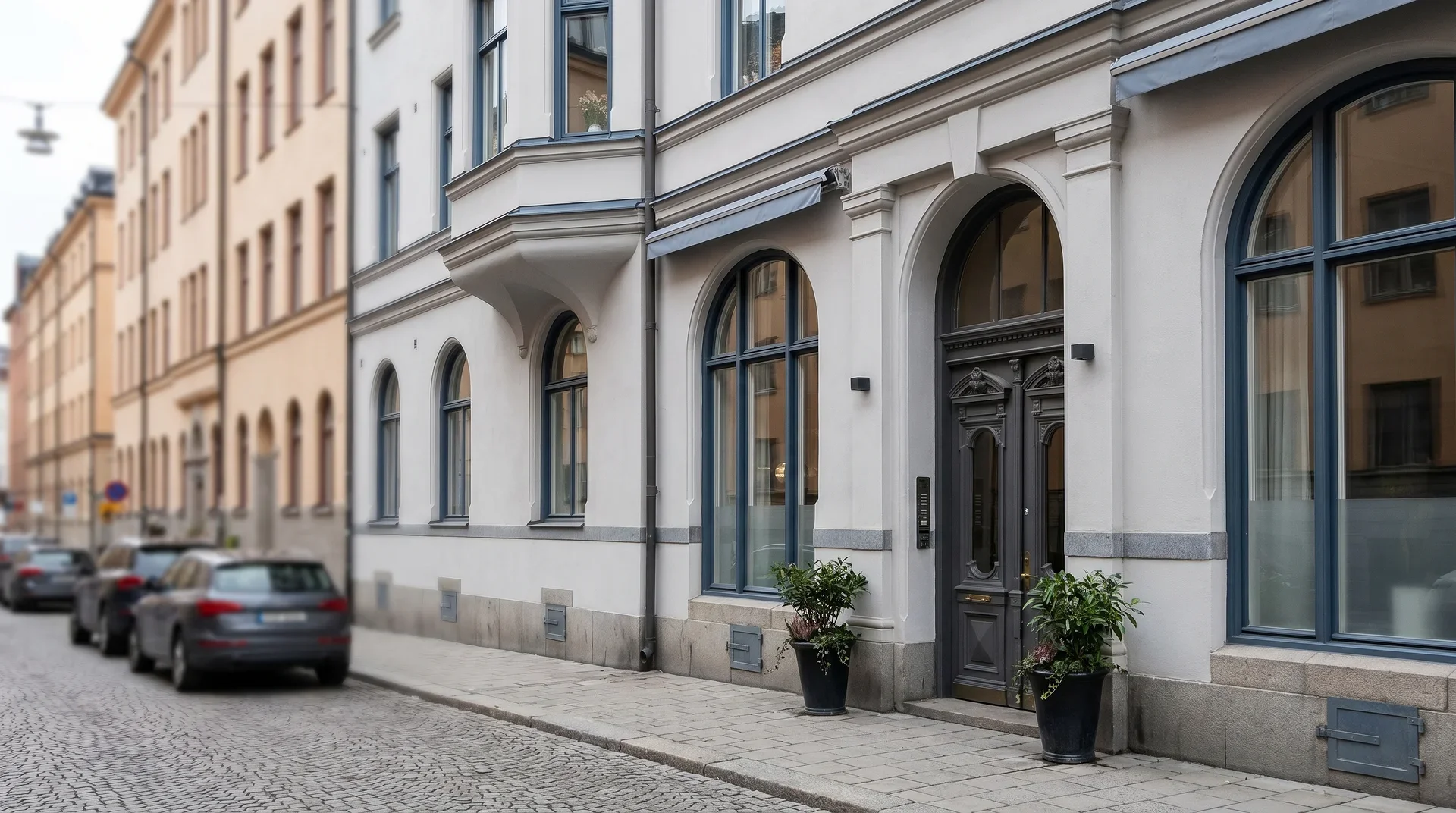 Klassisk vit stadsbyggnad med rundbågade fönster och entré vid kullerstenstorg.